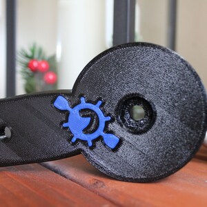 Op de afbeelding: Een zwarte 3D-geprinte peddelhouder met een blauw logo met een peddel en een wiel. De houder heeft vier gaten voor montage.