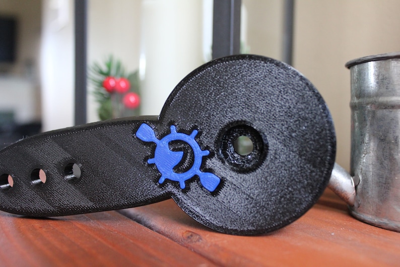 Op de afbeelding: Een zwarte 3D-geprinte peddelhouder met een blauw logo met een peddel en een wiel. De houder heeft vier gaten voor montage.