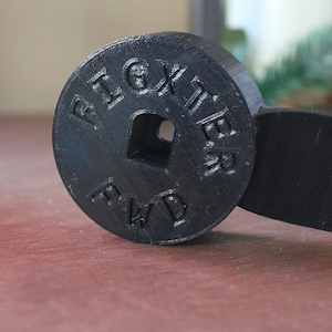 Op de afbeelding: Zwart plastic 3D-geprint onderdeel met de woorden "Fighter FWD" gegraveerd op een ronde schijf. De schijf is bevestigd aan een zwarte plastic arm met een gat in het midden.