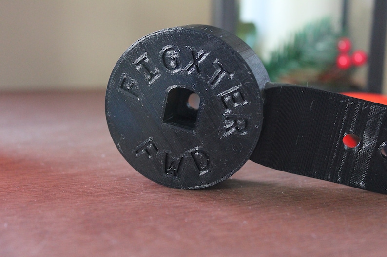 Op de afbeelding: Zwart plastic 3D-geprint onderdeel met de woorden "Fighter FWD" gegraveerd op een ronde schijf. De schijf is bevestigd aan een zwarte plastic arm met een gat in het midden.