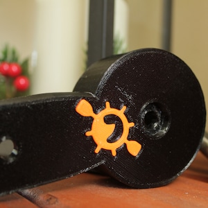 Op de afbeelding: Een zwart plastic peddel met een oranje logo met een gestileerde peddel en een wiel. Het logo is gecentreerd op de peddel.