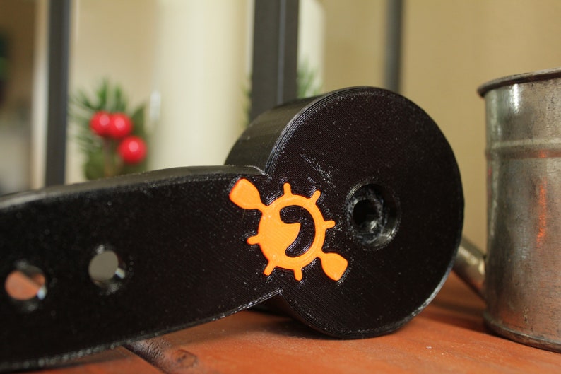 Op de afbeelding: Een zwart plastic peddel met een oranje logo met een gestileerde peddel en een wiel. Het logo is gecentreerd op de peddel.