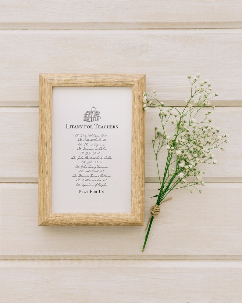 May include: A framed print with a light wood frame and a white background. The print features a litany for teachers with a stack of books and an apple on top. The text includes the names of saints and the phrase "Pray for Us".