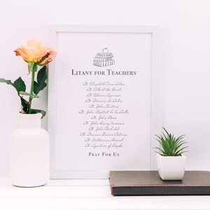 May include: A white framed print with a black and white illustration of a stack of books with an apple on top. The text "LITANY FOR TEACHERS" is at the top of the print. The print lists a litany of saints, including St. Elizabeth Ann Seton, St. Albert the Great, St. Thomas Aquinas, St. John Cantius, St. John Baptist de la Salle, St. John Bosco, St. John Henry Newman, St. John Paul II, St. Frances Xavier Cabrini, St. Catherine Drexel, and St. Josephine of Leghorn. The text "PRAY FOR US" is at the bottom of the print.