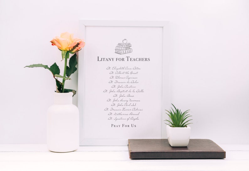 May include: A white framed print with a black and white illustration of a stack of books with an apple on top. The text "LITANY FOR TEACHERS" is at the top of the print. The print lists a litany of saints, including St. Elizabeth Ann Seton, St. Albert the Great, St. Thomas Aquinas, St. John Cantius, St. John Baptist de la Salle, St. John Bosco, St. John Henry Newman, St. John Paul II, St. Frances Xavier Cabrini, St. Catherine Drexel, and St. Josephine of Leghorn. The text "PRAY FOR US" is at the bottom of the print.