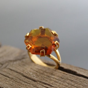 Puede incluir: Un anillo de oro con una gran piedra preciosa facetada de color ámbar. La piedra preciosa está engastada en una banda de oro con un diseño de caña dividida. El engaste del anillo tiene un diseño en forma de garra.