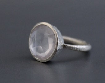 Puede incluir: Un anillo de plata con una gran piedra preciosa ovalada de color rosa pálido. El anillo tiene una banda texturizada y un engaste de bisel. El anillo está sobre un fondo gris oscuro. La piedra preciosa mide aproximadamente 1,5 cm de diámetro.