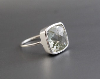 May include: A square-cut, light green gemstone ring with a silver band. The gemstone is faceted, reflecting light. The ring is set in a silver bezel, creating a modern and elegant design. The ring is a piece of jewelry.