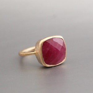 May include: A gold ring featuring a large, square-cut ruby gemstone. The ruby has a deep red color and is set in a gold bezel. The ring's band is a simple, thin gold band.