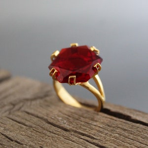 Pode incluir: Um anel de ouro com uma grande pedra preciosa vermelha escura facetada. A pedra preciosa é definida em uma configuração de garra de ouro, e o anel tem um design de haste dividida. O anel é exibido em uma superfície de madeira.