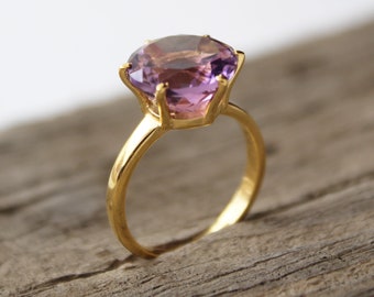 May include: A gold ring featuring a large, round, light purple gemstone. The ring's band and setting are a bright gold color, creating a striking contrast with the gemstone. The ring is set against a neutral background.