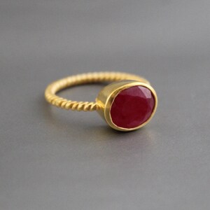 May include: A gold ring with a twisted band and a large, oval-shaped, deep red gemstone. The gemstone is set in a gold bezel, and the ring is displayed against a gray background. The ring is a piece of jewelry.