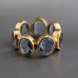 Puede incluir: Un anillo de oro con una banda de piedras preciosas azules. El anillo está hecho de un metal liso y brillante y las piedras preciosas están dispuestas en un patrón circular.