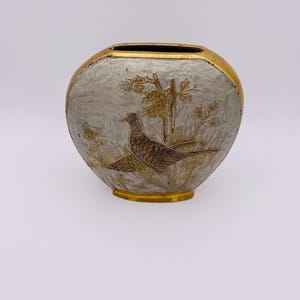 Op de afbeelding: Ovale vaas met een gouden rand en voet. De vaas heeft een gedetailleerde illustratie van een fazant in een veld, met gouden en bruine accenten. De achtergrond is een getextureerde, lichtgrijze kleur.