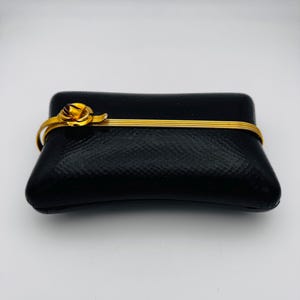 Può includere: Una pochette nera, strutturata, con una fascia color oro e una chiusura decorativa a forma di rosa. La pochette è rettangolare con bordi arrotondati. La fascia dorata avvolge il centro.