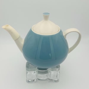 Op de afbeelding: Een blauw-witte keramische theepot met een afgeronde body en een gebogen tuit. De theepot heeft een wit deksel en handvat. Hij staat op een heldere, vierkante standaard. De theepot is lichtblauw.