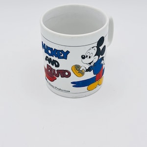Pode incluir: Uma caneca de cerâmica branca com um gráfico do Mickey Mouse segurando um sabonete amarelo. O texto diz Mickey and Pluto com Walt Disney Productions impresso abaixo. A caneca tem um corpo cilíndrico e uma alça no lado direito.