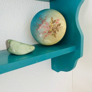 Könnte beinhalten: Ein blaues Holzregal mit zwei Gegenständen darauf. Das Regal hat eine dekorative Kante. Die Gegenstände sind eine runde, hellblaue und gelbe Schmuckschatulle mit einem Blumendesign und ein grüner, glatter, sichelförmiger Stein.