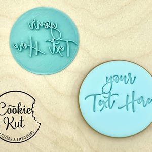 May include: Two round, light blue cookie cutters with the words "your Text Here" and "your Text" in a cursive font. The Cookie Kut logo is in the lower left corner.