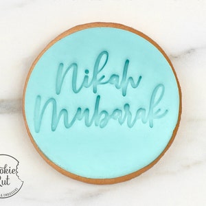 May include: A light blue round cookie with the words "Nikah Mubarak" written in white icing.