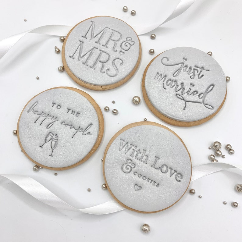 May include: Four round, silver-colored cookies with white icing and gray lettering. The cookies say "Mr. & Mrs.", "Just Married", "To the Happy Couple", and "With Love & Cookies".