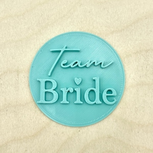 May include: A light blue round sticker with the text "Team Bride" in white lettering with a small heart symbol between the words.