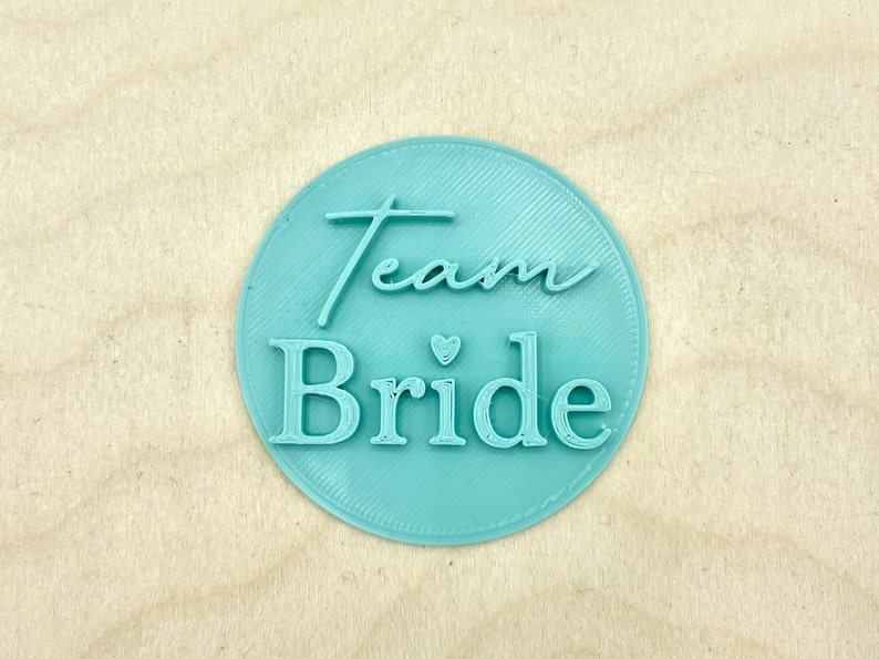May include: A light blue round sticker with the text "Team Bride" in white lettering with a small heart symbol between the words.