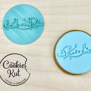 May include: A light blue circular cookie stamp with Arabic script, and a cookie with the same script. The stamp is next to a logo that says "Cookie Kut Cutters & Embossers".