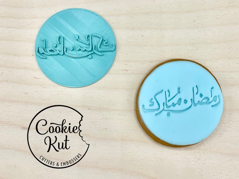 May include: A light blue circular cookie stamp with Arabic script, and a cookie with the same script. The stamp is next to a logo that says "Cookie Kut Cutters & Embossers".