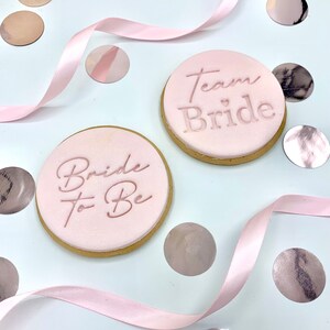 May include: Two pink sugar cookies decorated with pink icing and the text "Bride to Be" and "Team Bride" with a small heart symbol.