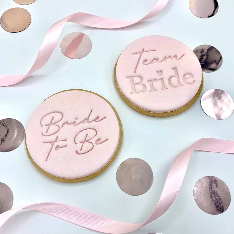 May include: Two pink sugar cookies decorated with pink icing and the text "Bride to Be" and "Team Bride" with a small heart symbol.