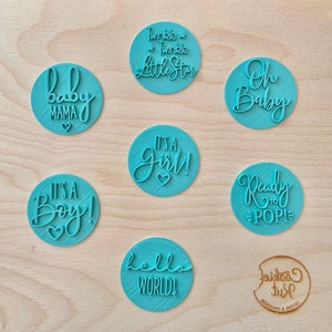 Op de afbeelding: Set van negen ronde blauwe koekjesuitstekers met verschillende babythema-zinnen, waaronder "Het is een jongen!" en "Twinkle Twinkle Little Star".