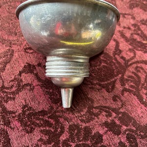 May include: A vintage metal funnel with a wide, bowl-shaped top and a narrow spout. The funnel is made of a shiny, silver-colored metal and has a threaded base. The funnel is likely used for pouring liquids or powders into containers.