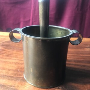 Könnte beinhalten: Ein Mörser und Stößel aus Messing mit zwei Griffen. Der Mörser ist zylindrisch und der Stößel ist ein langer, dünner Stab.