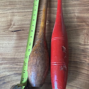 May include: Two wooden exercise clubs positioned next to a yellow measuring tape. One club has a natural brown wood finish, while the other is painted red. The measuring tape indicates a length of approximately 15 inches for the clubs.