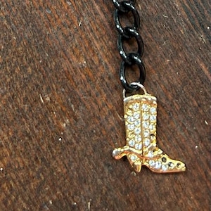 May include: A gold-colored cowboy boot charm with clear crystals hangs from a black chain. The charm is detailed with a gold-colored toe, heel, and top. The background is a wooden surface.
