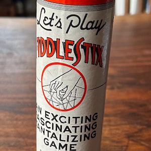 May include: A vintage cylindrical game container for "Fiddlestix." The red and white container features the text "Let's Play Fiddlestix" and "An Exciting, Fascinating, Tantalizing Game." The game is made by Plaza Mfg. Co. in New York.