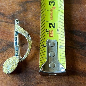 May include: A musical note brooch, embellished with yellow and clear rhinestones, is shown beside a yellow measuring tape. The brooch is about 3.8 cm long, featuring a detailed, sparkling design. The background is a wooden surface.