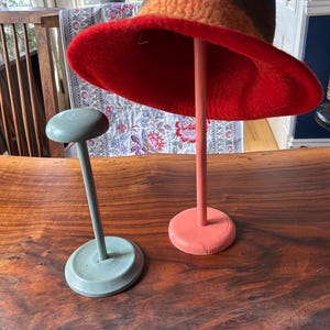 May include: A colorful wool hat with a wide red brim is displayed on a pink hat stand. A smaller, light green hat stand is next to it. The hat features bands of brown, orange, and red. The stands are on a wooden surface.