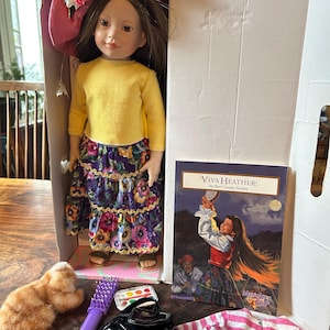 May include: A doll wearing a yellow long-sleeve shirt and a floral skirt, with a pink hat. Accessories include a stuffed animal, a brush, a backpack, a book titled "Viva Heather!", and a pink and white striped jacket.