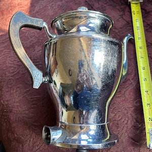 May include: A silver chrome coffee percolator with a curved handle and a spout. The percolator has a shiny, reflective surface.