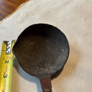 May include: A dark, antique-style ladle with a rounded bowl and a long handle. The ladle's bowl is approximately 3 inches in diameter. A yellow measuring tape is placed next to the ladle.