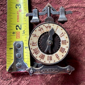 May include: A small, antique clock with a cream-colored face and Roman numerals. The clock is made of metal and has a decorative design. The clock measures approximately 2 inches in height.