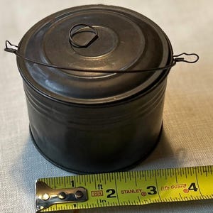 May include: A dark grey metal container with a lid and a wire handle. The container is cylindrical and measures approximately 10 cm in diameter and 7.6 cm in height. The lid has a small loop handle.