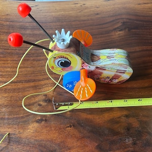 May include: A vintage pull-along bee-shaped toy with a yellow face, red antennae, and a patterned body. A yellow measuring tape is next to the toy, showing measurements in inches, equivalent to centimeters.