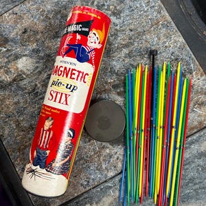 Op de afbeelding: Vintage "Magnetic Pick-Up Stix" spel. De rood-witte cilindrische container heeft illustraties van kinderen en een cartoon-elf. Een magnetische schijf en een bundel kleurrijke stokken zijn ook aanwezig.