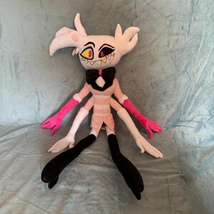 May include: A plush toy of a pink and white character with black legs and four arms. The character has a pink bow tie and a white and pink striped dress. The character has a yellow eye and a red mouth with sharp teeth.