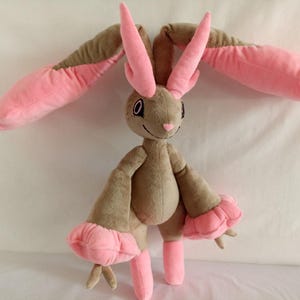 Puede incluir: Un juguete de peluche con forma de conejo con orejas largas de color rosa y pelaje marr&oacute;n. El juguete tiene una nariz rosa y grandes ojos negros.