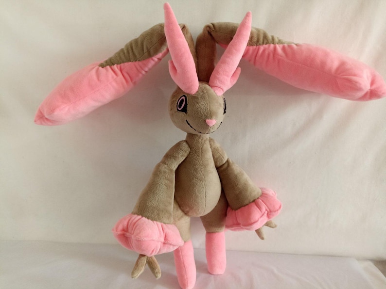 Puede incluir: Un juguete de peluche con forma de conejo con orejas largas de color rosa y pelaje marr&oacute;n. El juguete tiene una nariz rosa y grandes ojos negros.