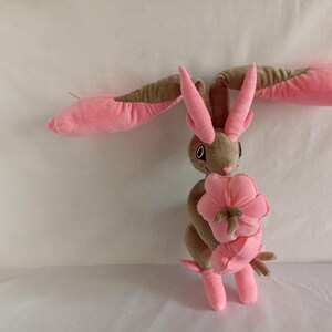 Puede incluir: Un conejo de peluche rosa y marr&oacute;n que sostiene una flor rosa. El conejo tiene orejas largas y un hocico largo.